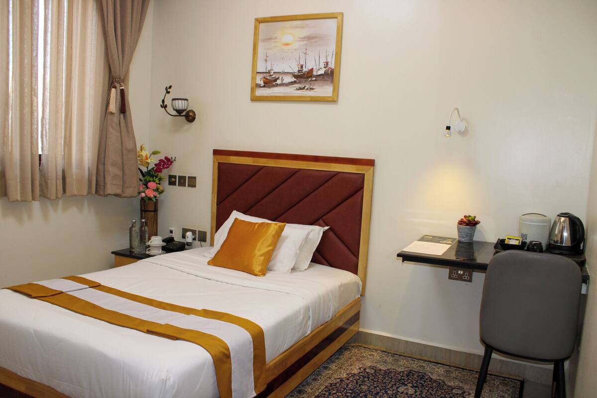 A hotel room with a bed, a desk with a chair, a wall-mounted lamp, a painting, and a window with curtains.
