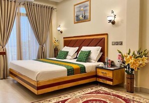 Quarto bem arrumado com cama de madeira, roupa de cama verde e branca, mesa de cabeceira com produtos de higiene pessoal e um vaso de flores.