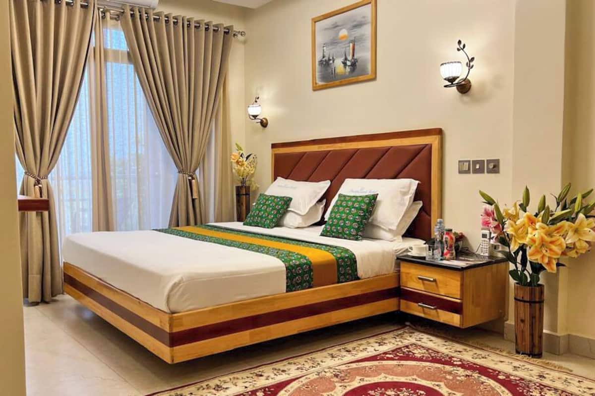 A hotel room with a large bed, beige curtains, a wooden headboard, a nightstand, a vase with flowers, and a painting on the wall.