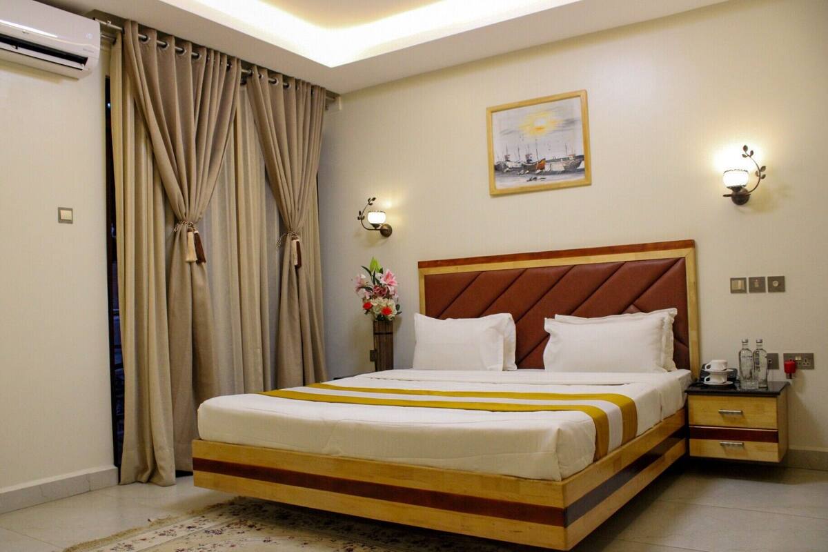 A hotel room with a bed, curtains, a wall-mounted lamp, a painting, and a nightstand with a cup and bottle.