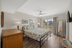 Un dormitorio con cama, lámparas de noche, ventilador de techo, cómoda de madera, ventana grande con vistas y puerta corrediza de cristal que da a un balcón.