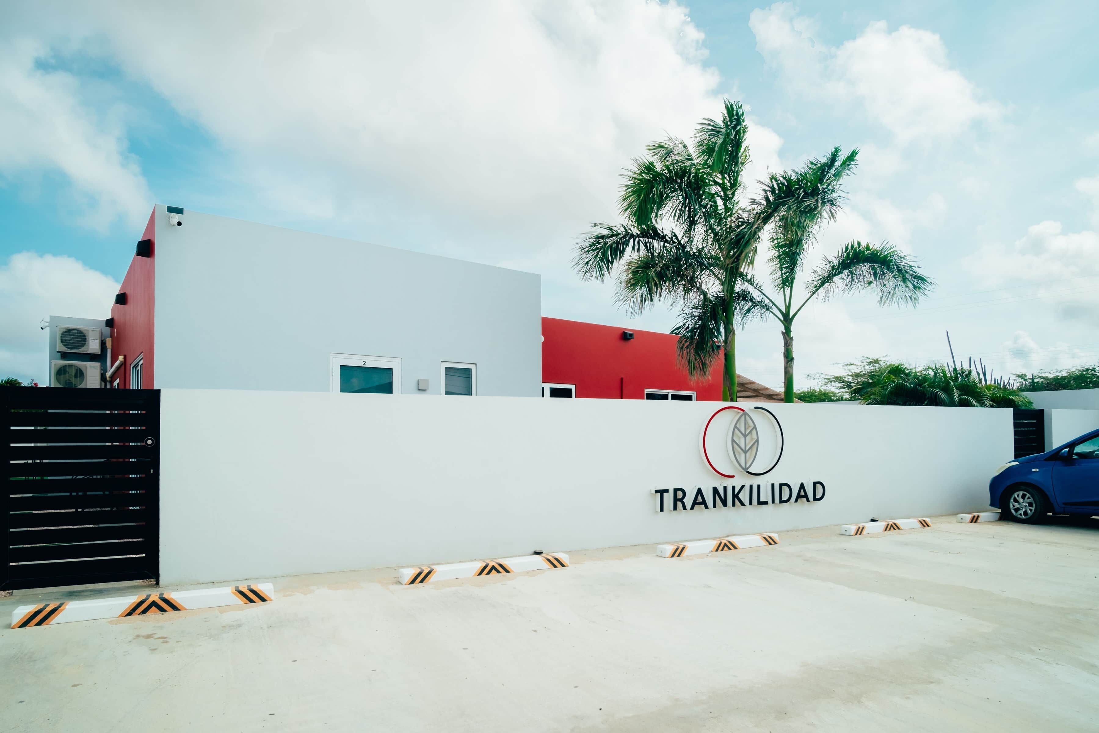 A gated property with a white wall and a red building, a palm tree, and a blue car parked in front.