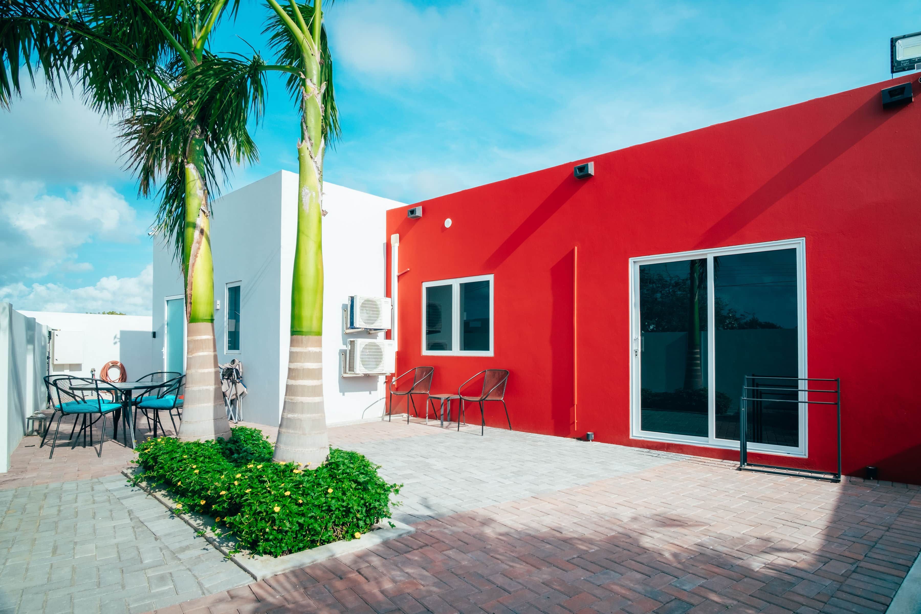A modern building with a red exterior, a patio area with outdoor furniture, and palm trees.