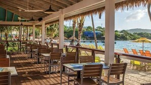 Un restaurante al aire libre con mesas y sillas de madera, techo de paja y vistas al mar y colinas.