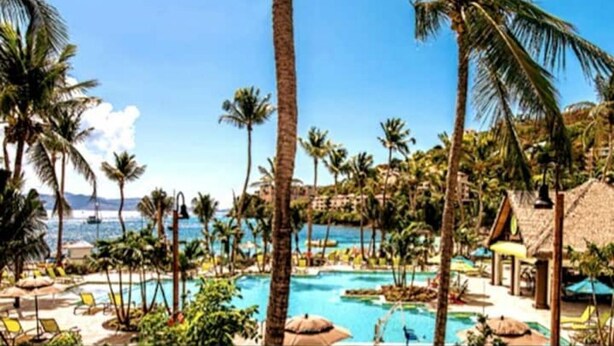Área de piscina en un resort, con palmeras, sillones de descanso y vista al océano.