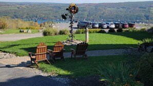 A parking area with a row of cars, a grassy area with a wooden chair, and a view of a lake and hills in the distance.