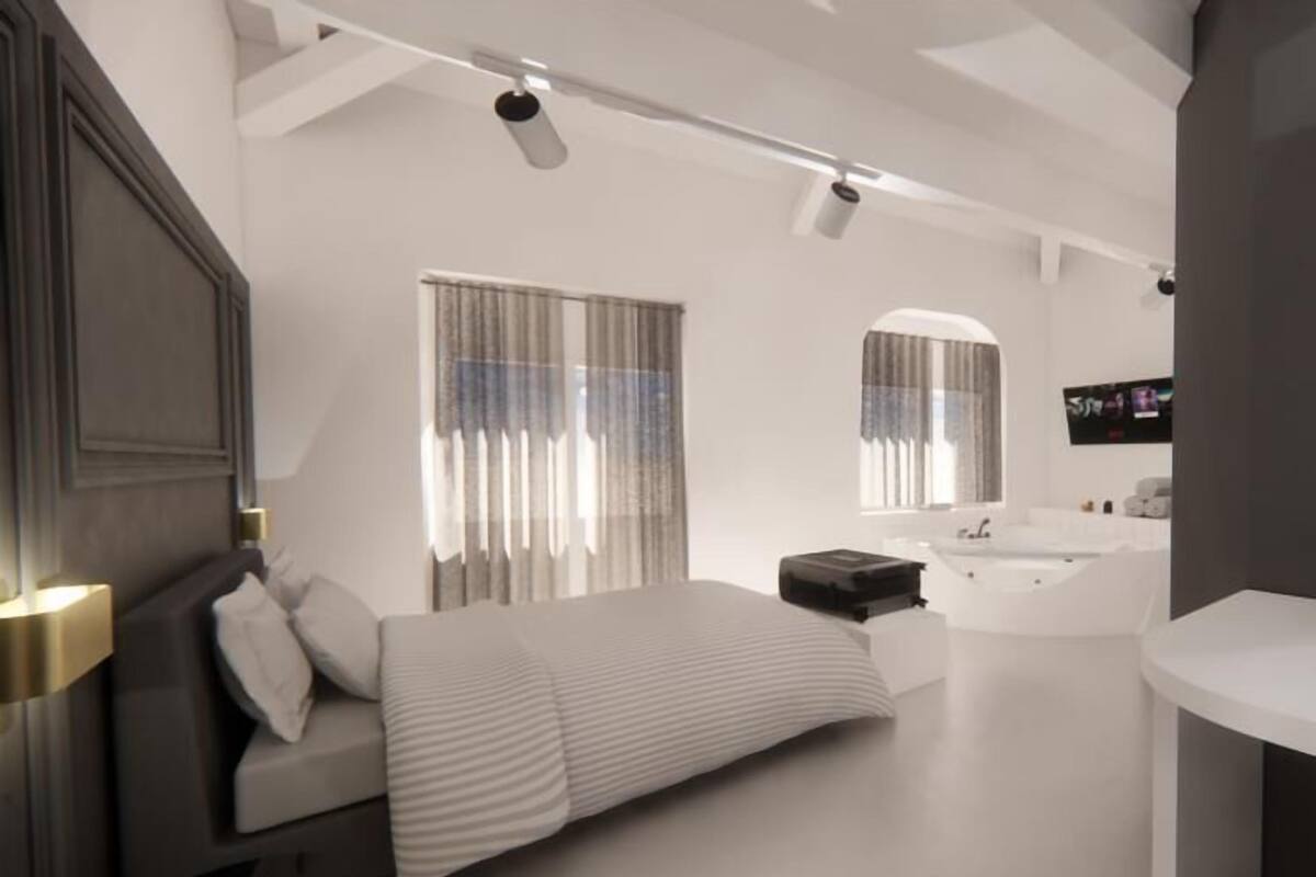 A modern bedroom with a large bed, a bedside table, a nightstand, a built-in bench, a large window with curtains, a wall-mounted TV, and a bathtub.