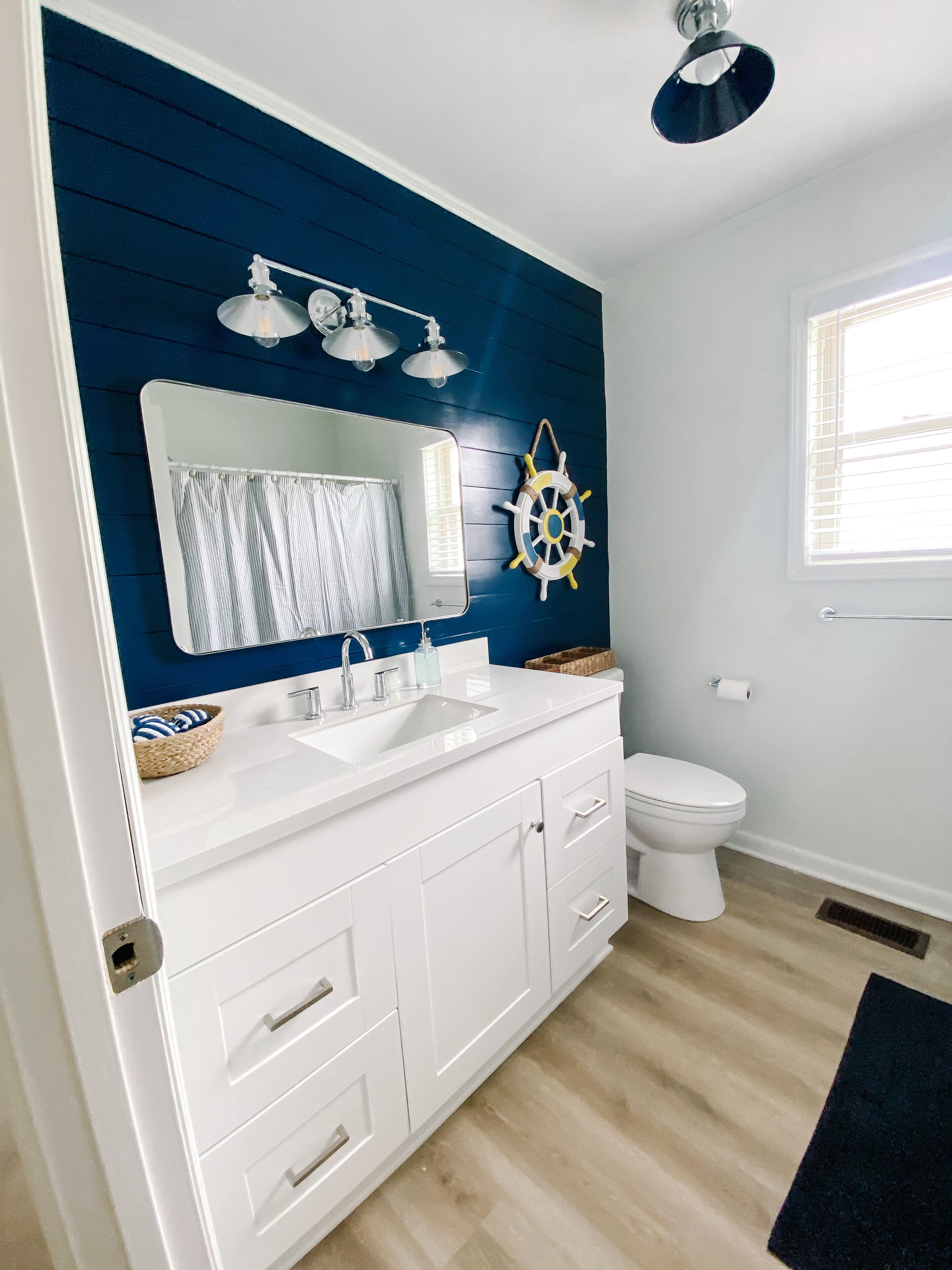 Un baño moderno con un tocador blanco, un espejo grande, una decoración de timón de barco y una ventana con persianas.