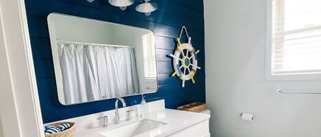 Un baño moderno con un tocador blanco, un espejo grande, una decoración de timón de barco y una ventana con persianas.