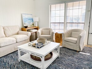 A living room with a white sofa, two armchairs, a coffee table with decorative items, a painting on the wall, and a window with blinds.