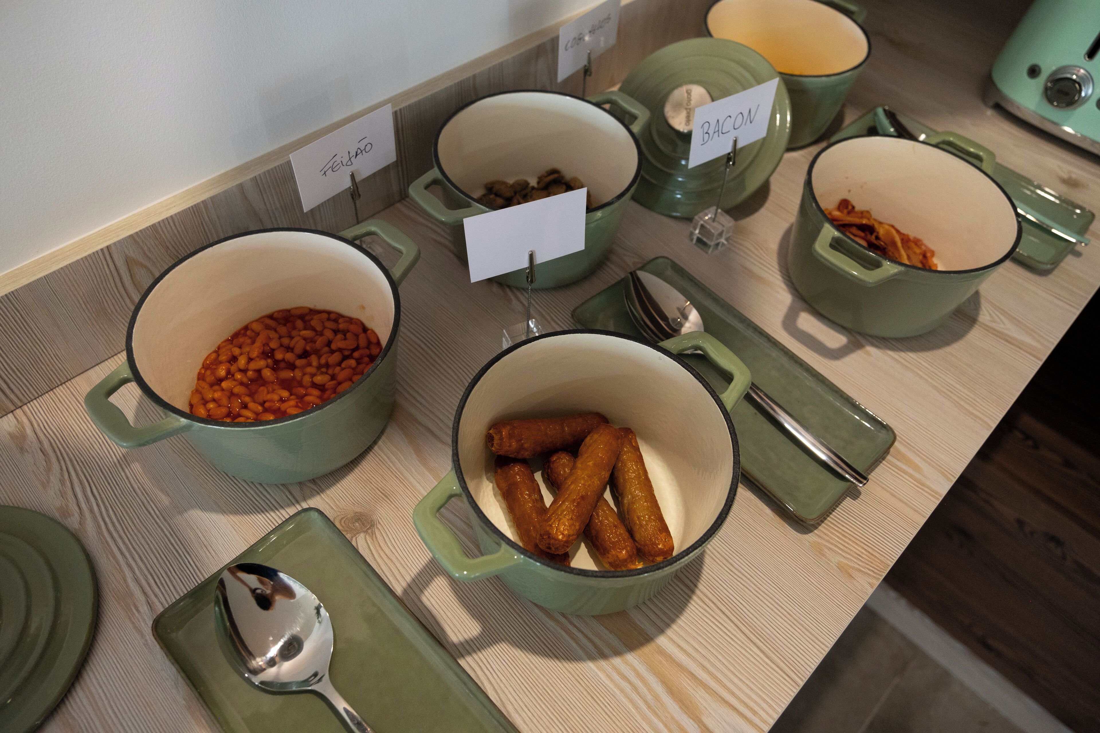 Een buffettafel met diverse gerechten in groene potten, waaronder bonen en worstjes, met aanduidingen zoals 'FEEDING' en 'BACON'.