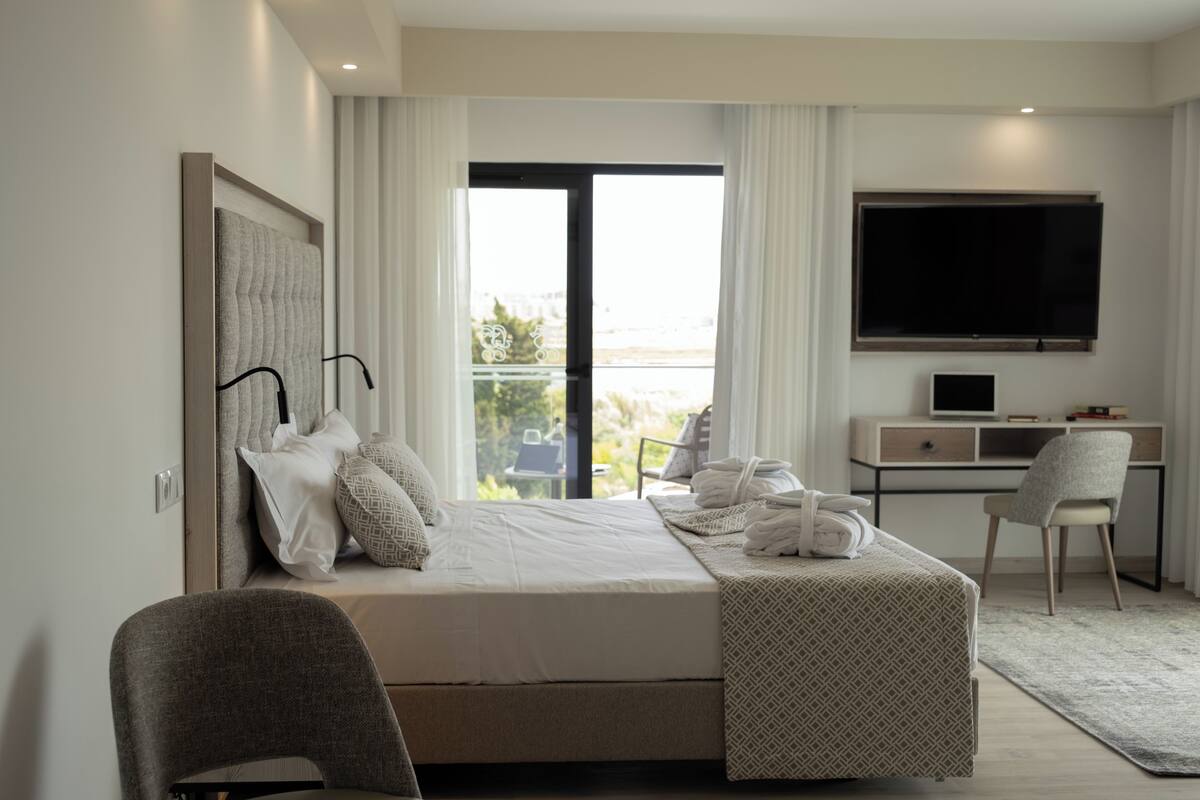 A modern hotel room with a large bed, a desk, a chair, a TV, and a balcony with a view.