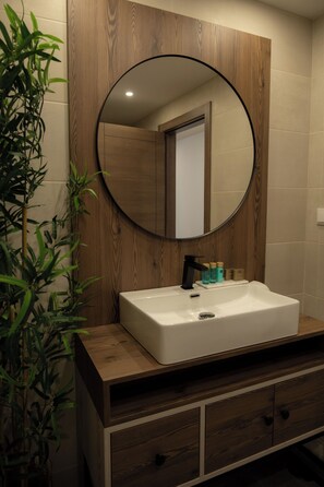 A bathroom with a wooden vanity, a white sink, a round mirror, and a potted plant.