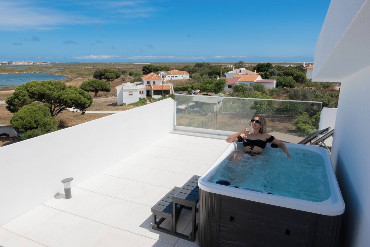 A person relaxing in a hot tub on a rooftop terrace with a view of the sea and surrounding buildings.