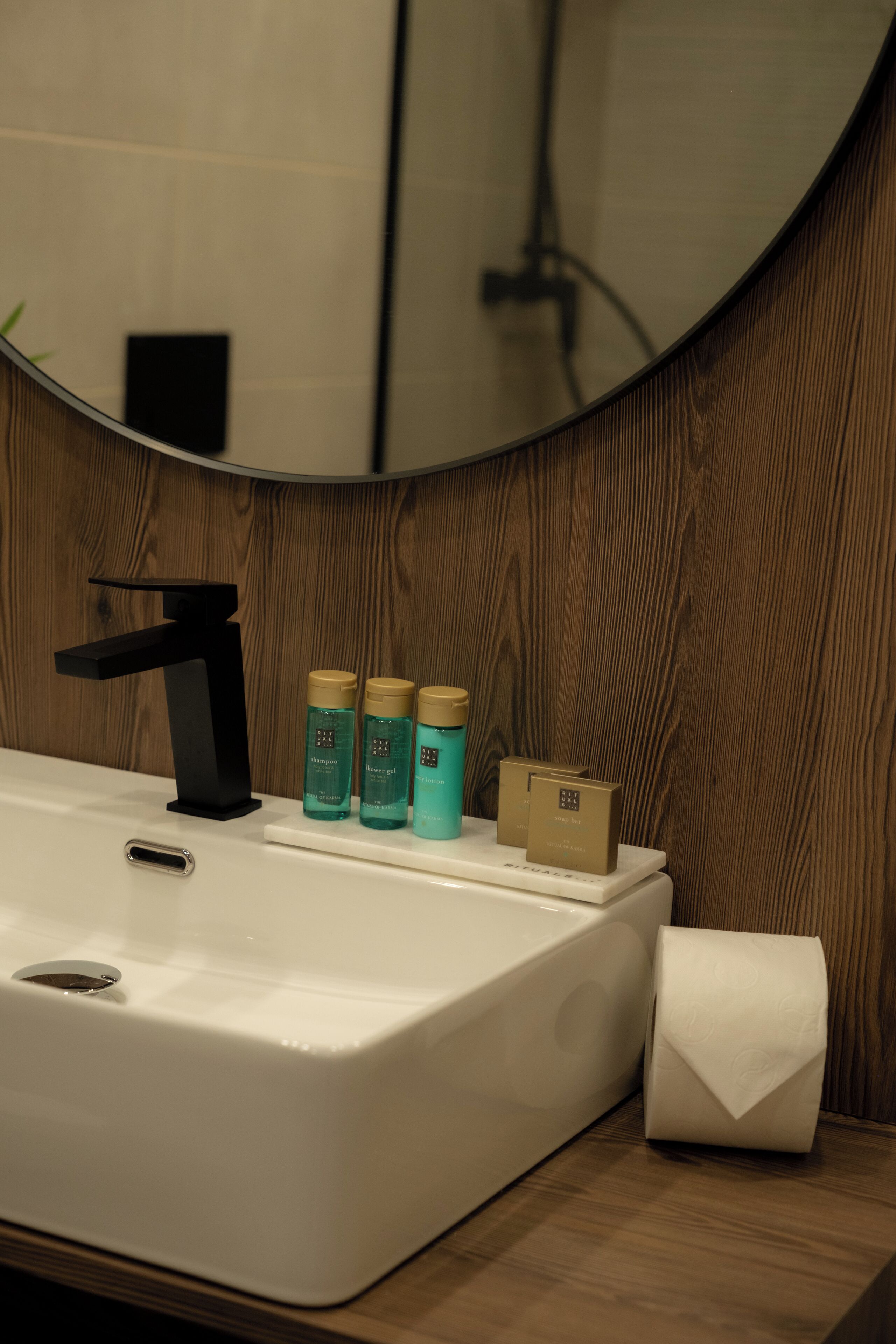 A bathroom sink with a black faucet, three toiletry bottles, a toilet paper roll, and a mirror reflecting a wooden wall.