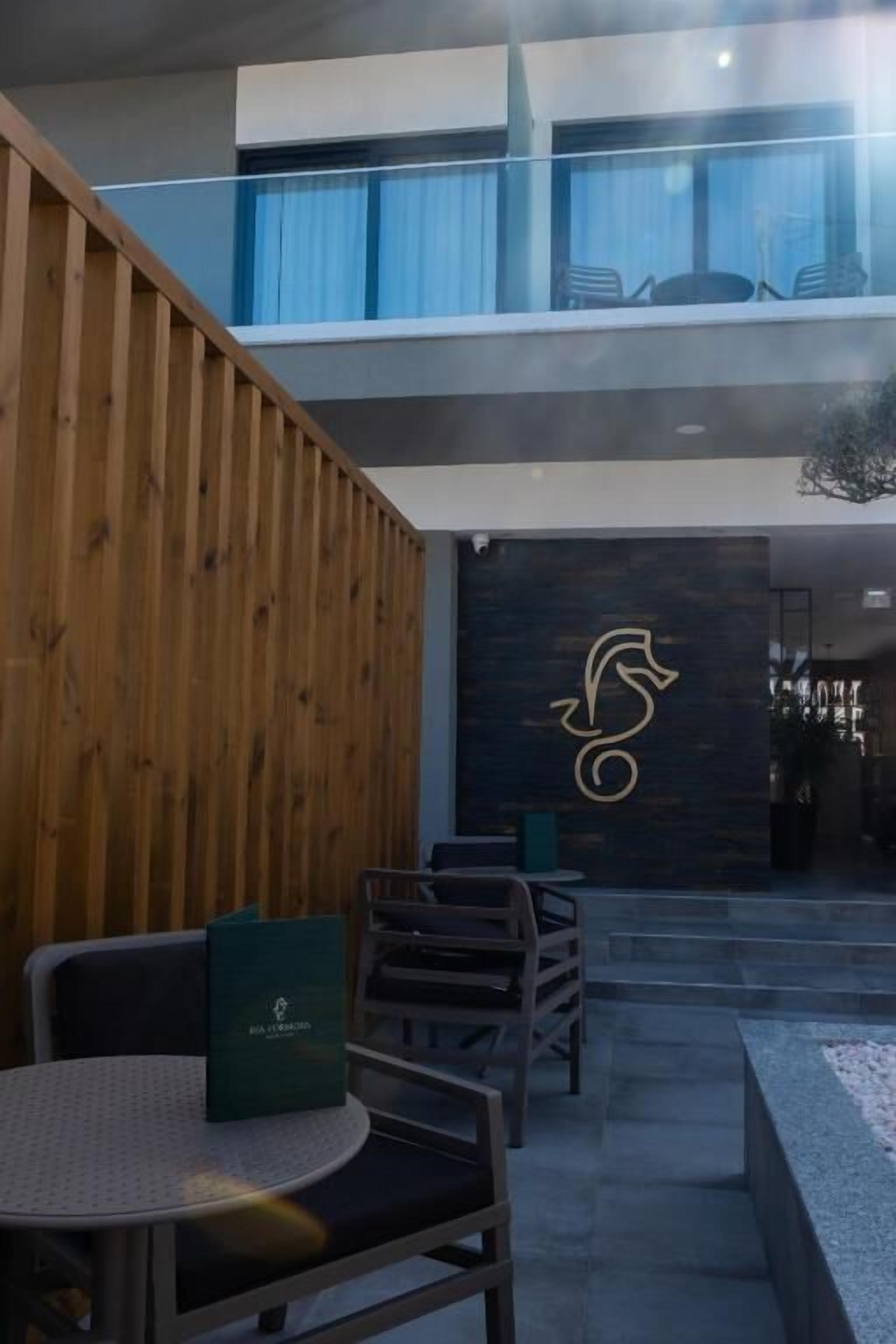 A modern outdoor seating area with wooden paneling, a stone wall with a logo, and a view of the upper floor.