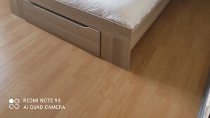 Ein ordentlich gemachtes Bett mit weißen Leinen und zwei zusammengerollten Handtüchern, ein Nachttisch aus Holz mit einer Lampe sowie eine Wand mit Muster.