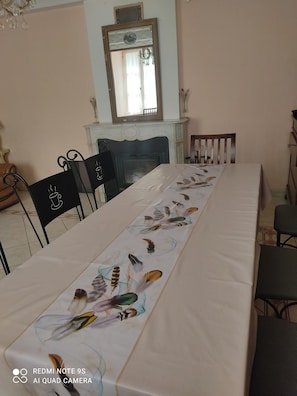 A dining area with a long table covered by a tablecloth with feather patterns, black chairs, a fireplace, and a mirror on the wall.