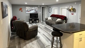 Un espace de vie compact comprenant un canapé, un fauteuil inclinable, un téléviseur et un coin cuisine.