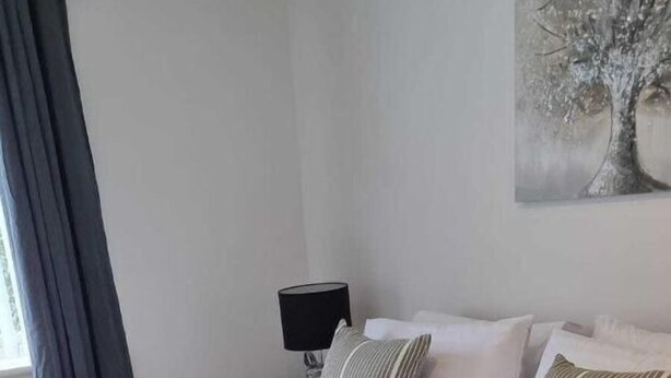 Una cama bien hecha con una manta y almohadas verdes, una lámpara de noche y un cuadro de un árbol en la pared.