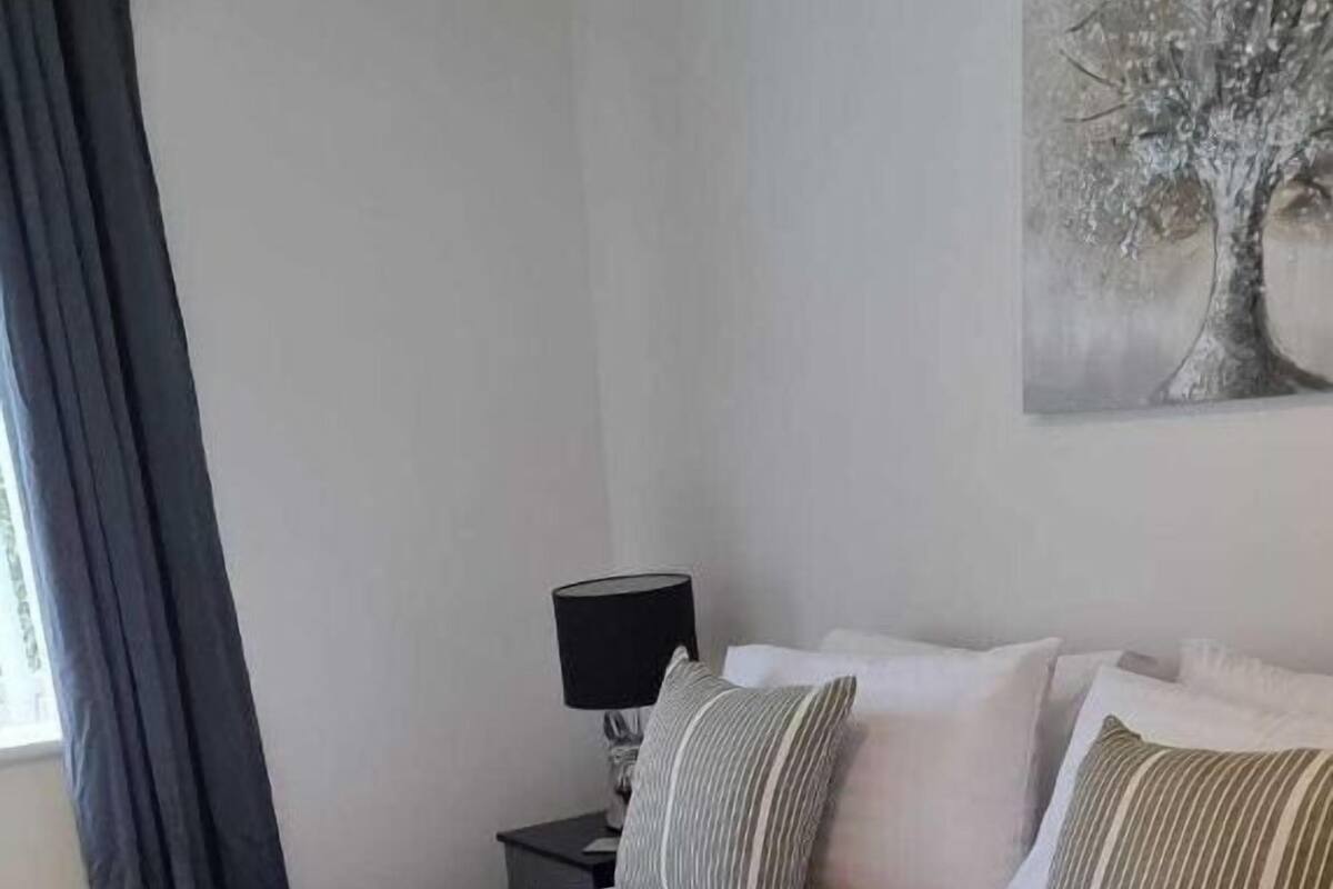 Una cama bien hecha con una manta y almohadas verdes, una lámpara de noche y un cuadro de un árbol en la pared.