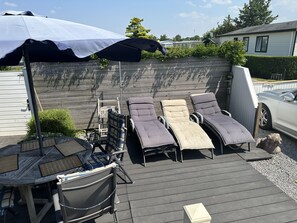 Eine Terrasse mit Liegestühlen und einem Tisch, ein Sonnenschirm und ein Haus im Hintergrund.