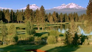 Um campo de golfe com um lago, árvores e montanhas cobertas de neve ao fundo.