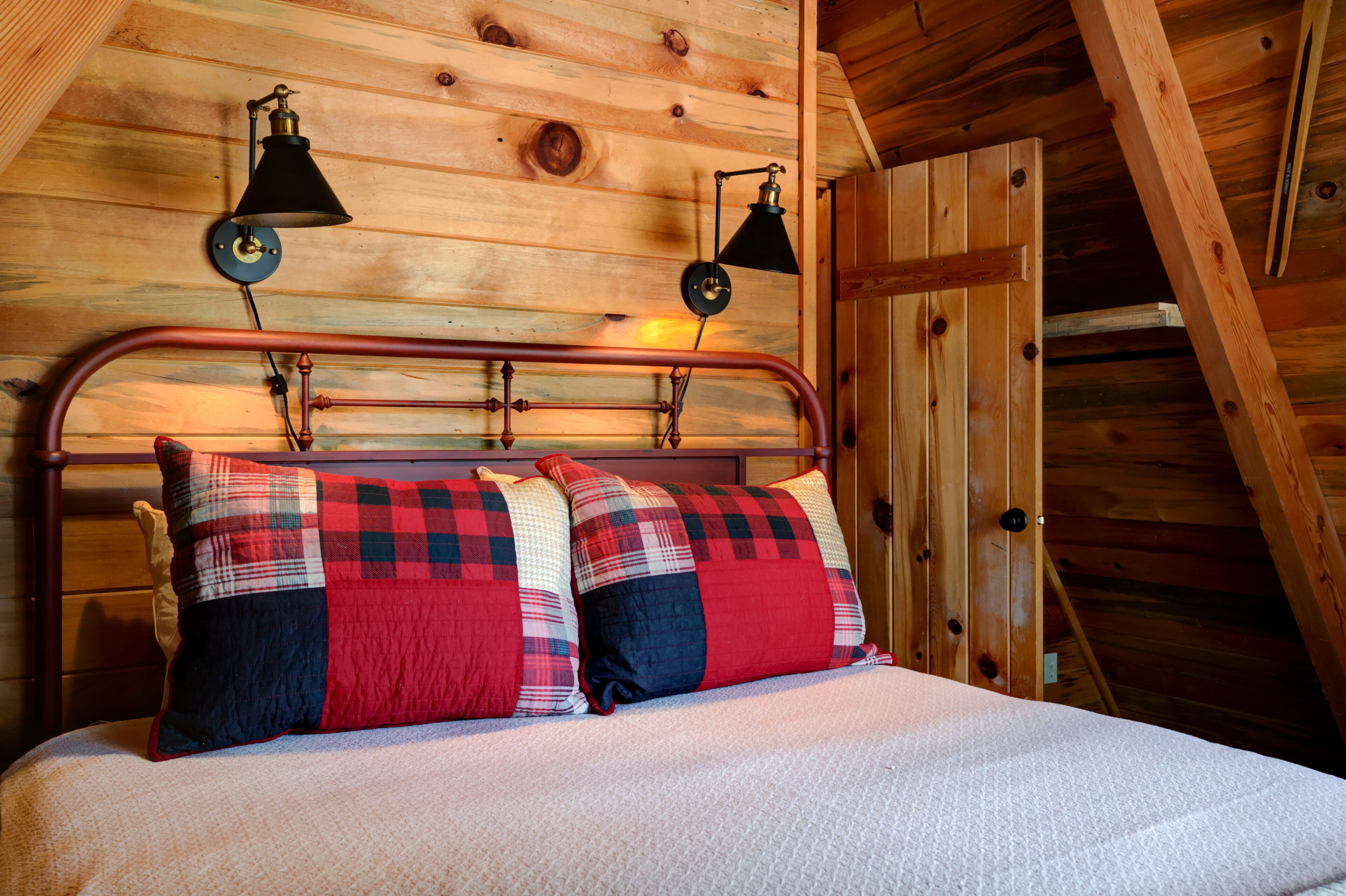 A wooden cabin bedroom with a metal bed frame, plaid bedding, and two wall-mounted lamps.