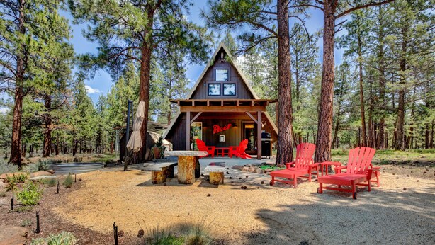 Uma cabana com uma placa de 'Beer', rodeada por árvores, com área de estar ao ar livre e chão de cascalho.