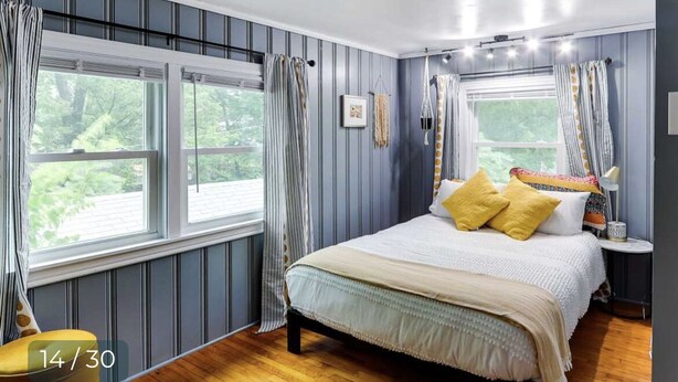 Un dormitorio con una cama grande, dos ventanas con cortinas, una mesita de noche con lámpara y vistas a la vegetación exterior.