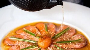 Un piatto con fette sottili di pesce disposte a cerchio, guarnito con erbe aromatiche verdi e semi di sesamo, e salsa che viene versata sopra.