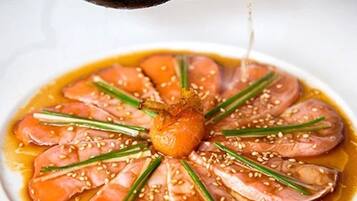 Un plato con láminas finas de pescado dispuestas en forma circular, adornado con hierbas verdes y semillas de sésamo, y salsa vertida encima.