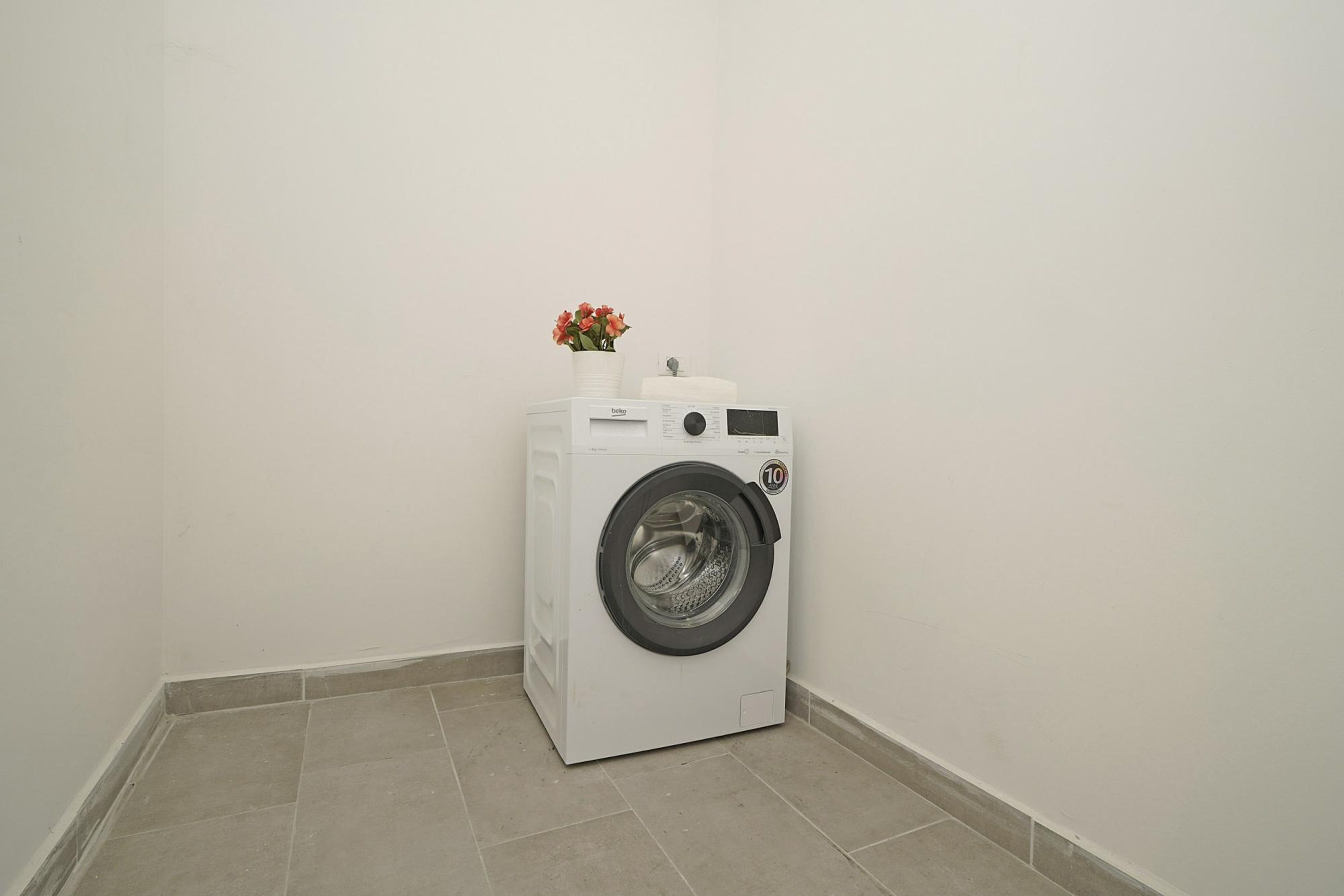 A compact, white washing machine in a corner with a small flower vase on top.