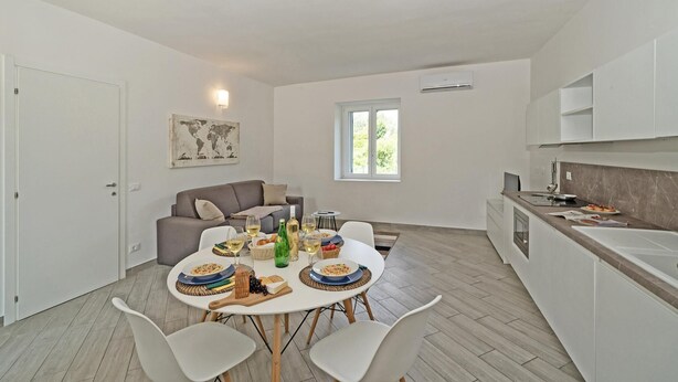 A modern kitchen with white cabinets, a dining table set for four, and a sofa in the living area. - San Carlo Borromeo 1 by Wonderful Italy (Soiano del Lago)