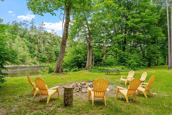 Un terrain de camping avec un foyer entouré de chaises Adirondack, une table de pique-nique et une rivière en arrière-plan.