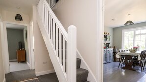 Un escalier avec une rampe blanche menant à un palier, un coin cuisine avec une table et des chaises, et un couloir agrémenté d’un tapis à motifs.