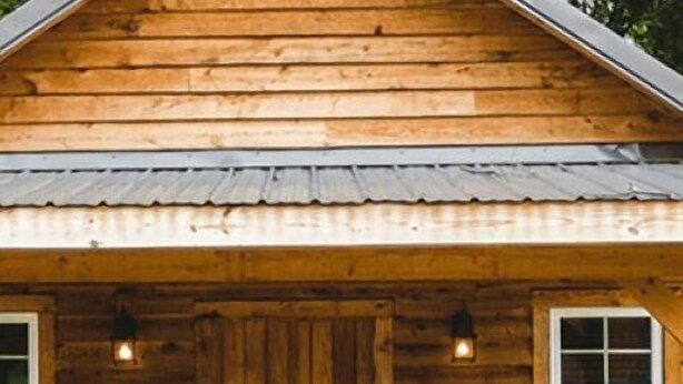 Una cabaña de madera con puerta principal, dos ventanas y dos sillas afuera.