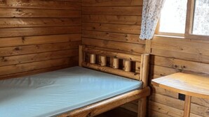 Una pequeña habitación de cabaña de madera con una cama individual, una mesita y una ventana con cortina de encaje.