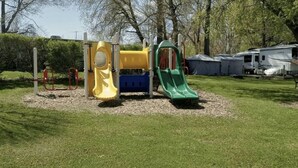 Un parque infantil con un tobogán amarillo, una estructura azul para escalar y un tobogán verde, rodeado de césped y árboles.