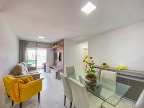 A modern living room with a glass dining table, a sofa with yellow cushions, a flat-screen TV mounted on a wooden panel, and a balcony with a view.