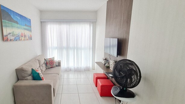 A living room with a sofa, red ottoman, TV, and a fan. - Flats no Muro Alto Condomínio Club (Ipojuca)