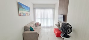 A living room with a sofa, red ottoman, TV, and a fan.