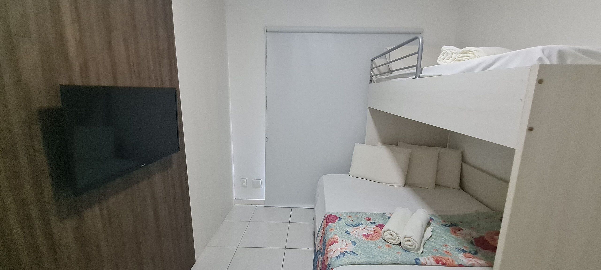 Uma cama beliche com uma televisão fixada na parede, uma porta e um chão ladrilhado.