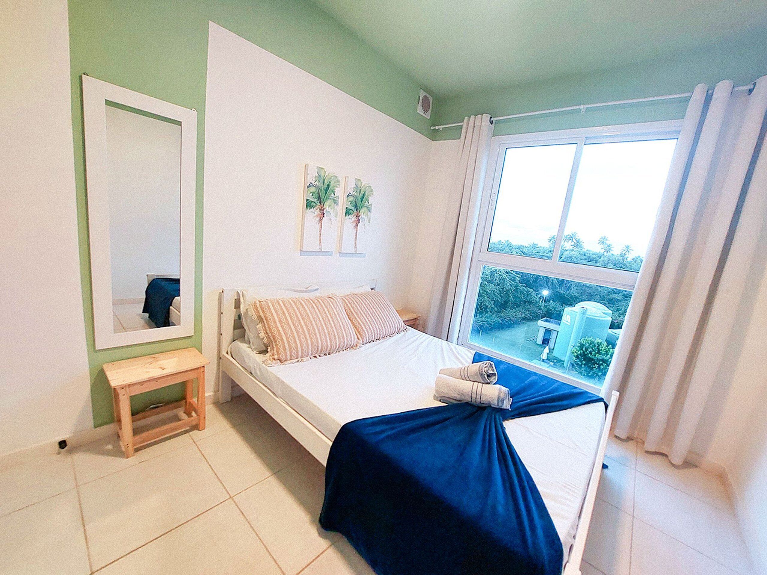 A bedroom with a bed, a small bedside table, a window with curtains, and a mirror on the wall.