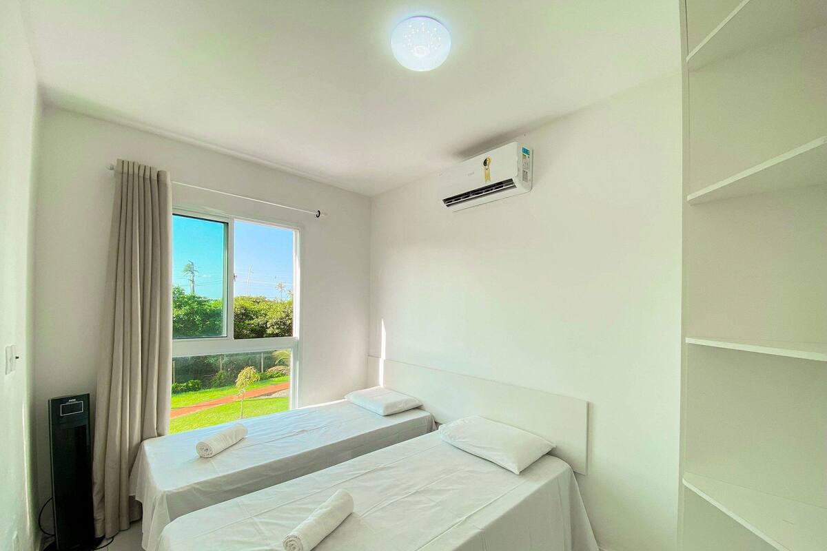 A hotel room with two beds, an air conditioning unit, a window with curtains, and a view of greenery outside.