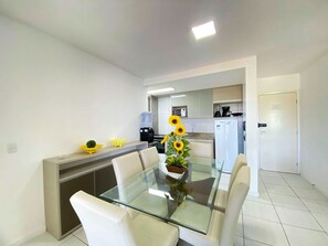 A modern dining area with a glass table, white chairs, and a kitchen in the background.