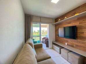 A modern living room with a sofa, a flat-screen TV mounted on a wooden wall, and a sliding glass door leading to an outdoor area. - Flats no Muro Alto Condomínio Club (Ipojuca)