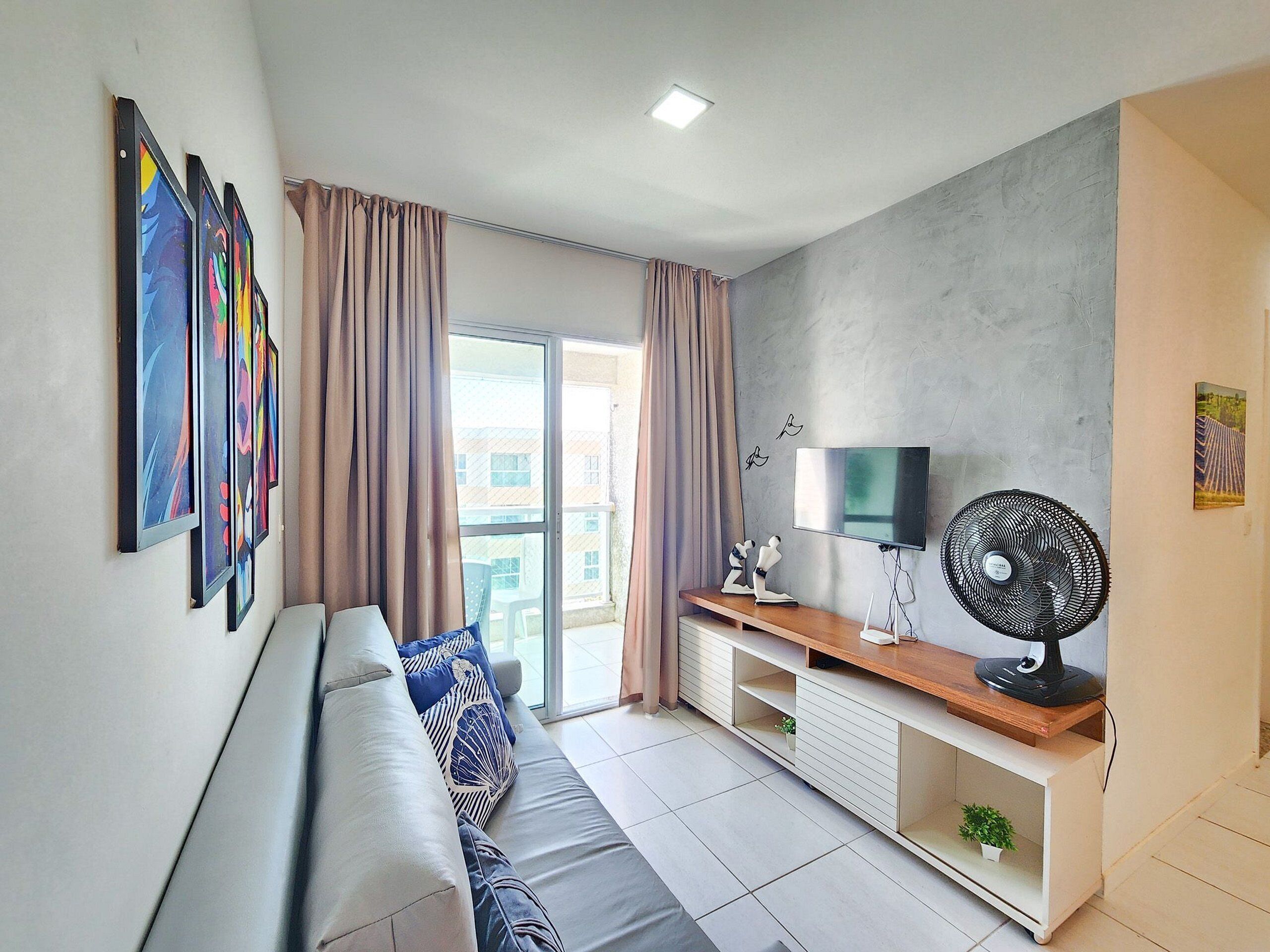 A modern living room with a sofa, TV, and fan, featuring artwork on the wall and a balcony view.
