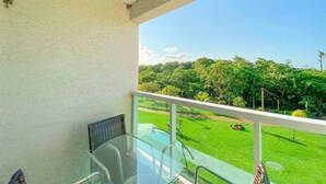A balcony with a glass table and chairs, a white railing, and a view of a well-maintained lawn and trees. - Flats no Muro Alto Condomínio Club (Ipojuca)