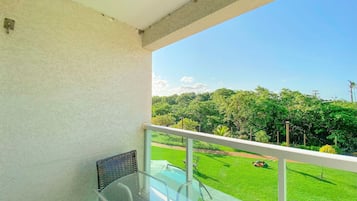 A balcony with a glass table and chairs, a white railing, and a view of a well-maintained lawn and trees.
